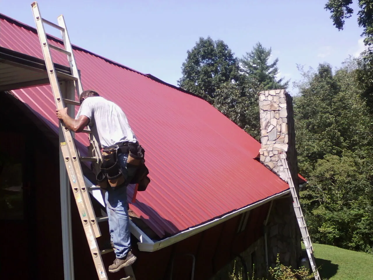 Roofer on a ladder beside a red metal roof and stone chimney — Soffit & Fascia Repair by Roofing Brothers in Harwich